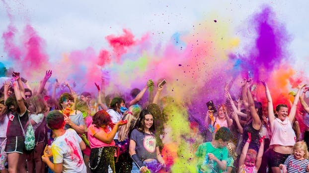 A joyful crowd covered in vivid colors celebrates a festival outdoors with flying powder.